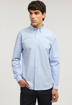 Junger Mann, der ein hellblaues Hemd mit dezentem Karo-Muster trägt, mit einer Hand in der Tasche stehend und zur Seite blickend.