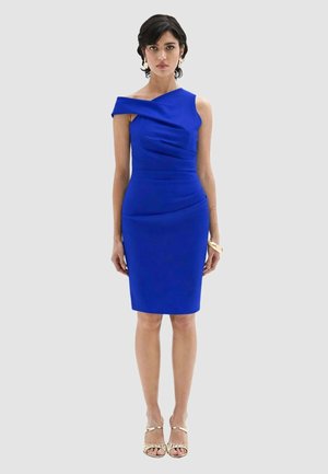 Donna in piedi con un vestito aderente blu cobalto monospalla al ginocchio, orecchini dorati, braccialetto e sandali con cinturini su uno sfondo grigio.