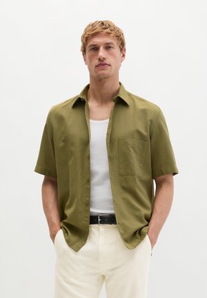 Blonde man draagt een open olijfgroen kort mouwen overhemd over een wit hemd, met crèmekleurige broek en zwarte riem, staand met handen in de zakken.