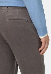 Pantalones de pana grises con textura de canalé vertical, que cuentan con un bolsillo con cremallera y una etiqueta de logo minimalista en la parte trasera, llevados con un suéter azul claro.