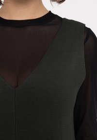 Haut tricoté texturé vert foncé avec un décolleté en V profond, superposé sur une chemise noire à manches longues transparente. Détail de couture visible au centre.
