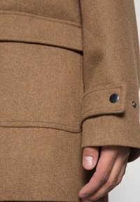 Wollen blendjas in bruin, met een opvallende kraag, decoratieve stiksels en een knoopdetail op de mouwen. Zachte textuur en gestructureerd ontwerp.
