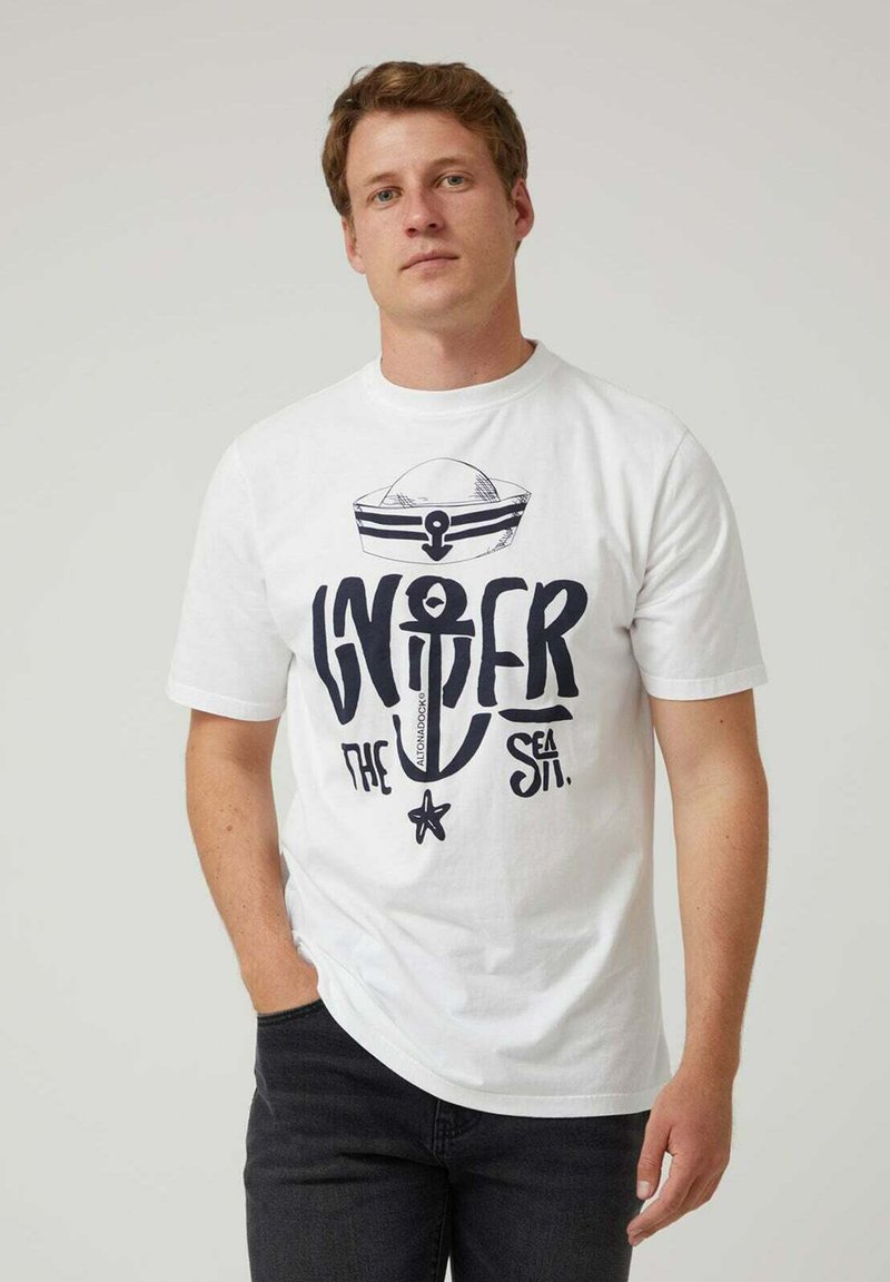 Camisa de algodón blanca con un audaz diseño gráfico en azul marino que presenta un ancla, un barco y el texto "Bajo el Mar". Cuello redondo y mangas cortas.