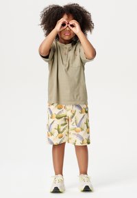 Chemise olive verte à manches courtes avec trois boutons en bois, associée à un short beige orné de motifs colorés de fruits et de feuilles tropicales. Chaussures blanches.