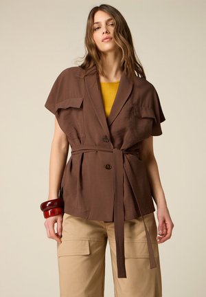 Vrouw met een bruine korte mouwen jas met riem over een mosterdkleurige top, beige cargobroek en grote rode armbanden aan haar linkerpols.