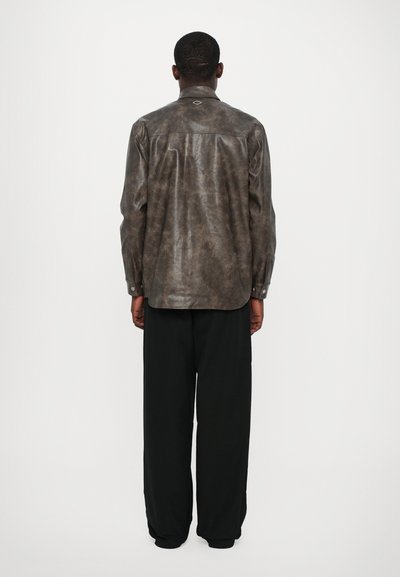 Chemise en cuir marron avec une finition lisse et texturée. Elle possède des manches longues, un col pointu et un petit détail brodé à l'arrière.