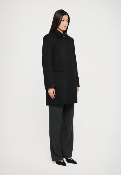 Black wool overcoat with a straight silhouette, button closure, and side pockets, paired with gray pinstriped trousers and black heels.