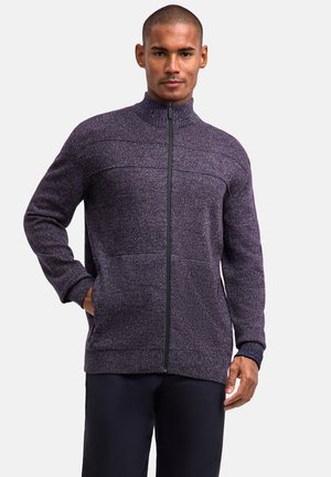 Hombre con un jersey con cremallera texturizado de color púrpura con bolsillos y pantalones negros, de pie con una mano en el bolsillo sobre un fondo sencillo.