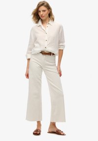 Witte linnen overhemd met knopen en lichtcrème wijde broek met rafelige zoom. Een gevlochten bruine riem en bruine slidesandalen maken de outfit compleet.