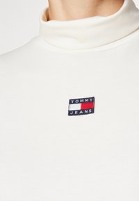 White turtleneck sweater with ribbed texture. Features a small, embroidered rectangular logo in navy, red, and white on the chest.