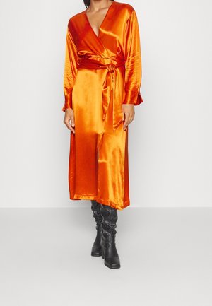 Vrouw die een glanzende oranje wikkeljurk met lange mouwen en een riem draagt, gecombineerd met zwarte kniehoge laarzen, staand tegen een witte achtergrond.
