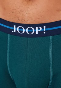 Pánské boxerky v tmavě zelené látce s strukturovaným povrchem. Mají námořnicky modrý pas s nápisem "JOOP!" a světle modrý pruh.