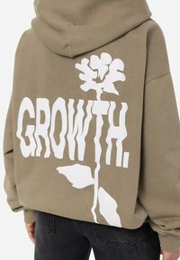 Felpa verde oliva con testo bianco audace "GROWTH." e un design floreale sul retro. Tessuto morbido con una vestibilità comoda e cappuccio.