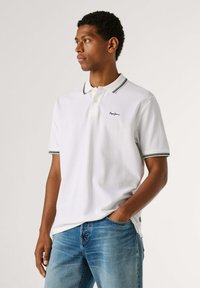 Weißes Polohemd mit klassischem Kragen, schwarz-navy gestreiften Akzenten an Kragen und Ärmeln, mit Logo auf der linken Brust. Legere blaue Jeans.