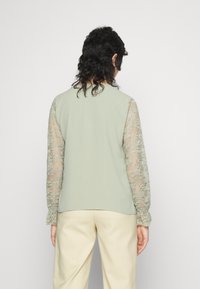 Top verde claro com mangas longas de renda transparente, apresentando um corpo de tecido suave e punhos franzidos. Sem padrões visíveis ou detalhes adicionais.