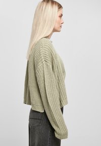 Ljusgrön stickad tröja med en cropped design, vida ärmar och ribbade muddar. Buren över en mörkgrå kjol, som visar upp textur och stygnmönster.