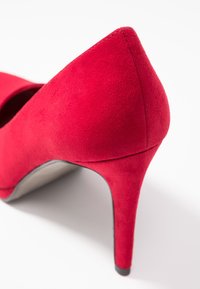 Červená sametová vysoká bota na podpatku s špičatou špičkou a štíhlým jehlovým podpatkem. Vyznačuje se hladkou texturou a čistým designem bez viditelného kování.