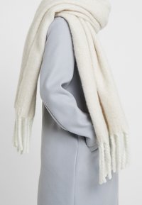 Écharpe en laine blanche à la texture douce, aux extrémités frangées, drapée sur un manteau gris clair. L'écharpe semble épaisse et confortable.