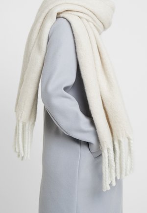 Écharpe en laine blanche à la texture douce, aux extrémités frangées, drapée sur un manteau gris clair. L'écharpe semble épaisse et confortable.