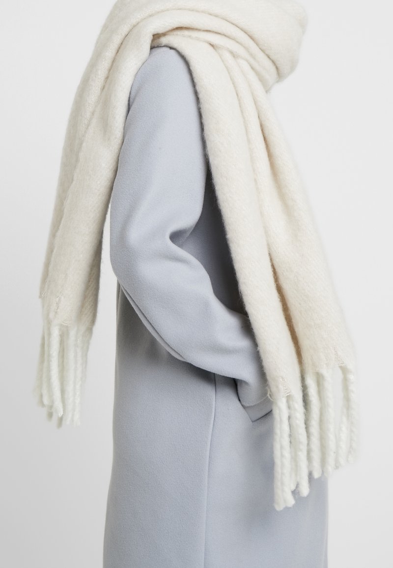 Écharpe en laine blanche à la texture douce, aux extrémités frangées, drapée sur un manteau gris clair. L'écharpe semble épaisse et confortable.