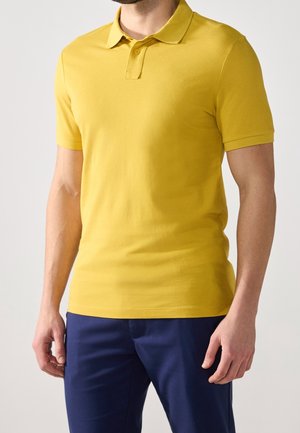 Man met een mosterdgele polo met korte mouwen en donkerblauwe broek, staand voor een effen lichte achtergrond.