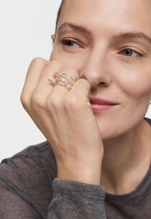 Mujer apoyando la barbilla en la mano, con un anillo plateado de varias bandas, sonriendo suavemente y mirando hacia un lado sobre un fondo liso.