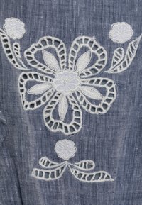 Denimová tkanina má bílé květinové vyšívání s krajkovými akcenty, včetně velké centrální květiny a menších květů, na texturovaném modrém pozadí.