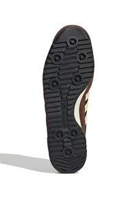 Brun idrottssko sula med svart gummitractionsmönster, centrala spår och cirkulära greppaccenter; texturerad yta för förbättrat grepp.