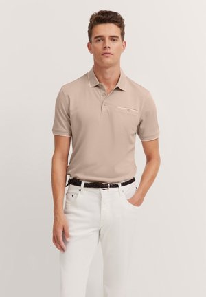 Beiger Poloshirt mit einer mit Knopf versehenen Brusttasche, kurzen Ärmeln und einem kontrastierenden Kragen. Kombiniert mit weißen Hosen und einem geflochtenen schwarzen Gürtel.