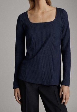 Femme portant un haut ajusté en maille côtelée bleu marine à manches longues avec un col carré et un pantalon noir, sur un fond uni.