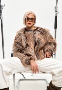 Chaqueta de piel sintética en tonos marrones con un diseño voluminoso y cuello alto. La modelo lleva gafas de sol oversized y pantalones crema.