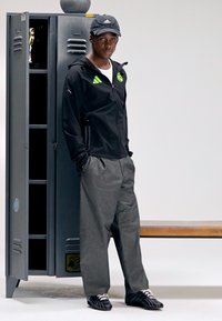 Athletic jacket in black with green accents, gray loose trousers, and black shoes with white laces; stands beside a gray locker.