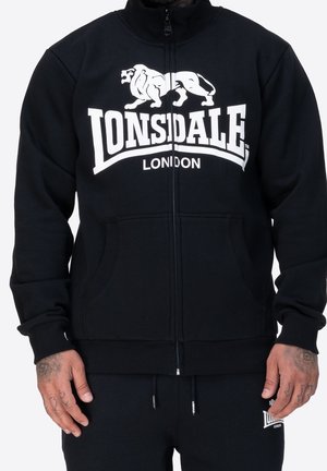 Osoba nosząca czarną bluzę z kapturem na zamek z białym logo „Lonsdale London” oraz grafiką lwa na piersi, w połączeniu z czarnymi spodniami.