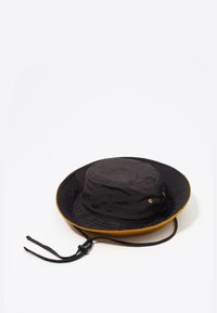 Sombrero de cubo de ala ancha en negro y mostaza con correa ajustable para el mentón, ojales de metal para ventilación, dispuesto sobre un fondo blanco.