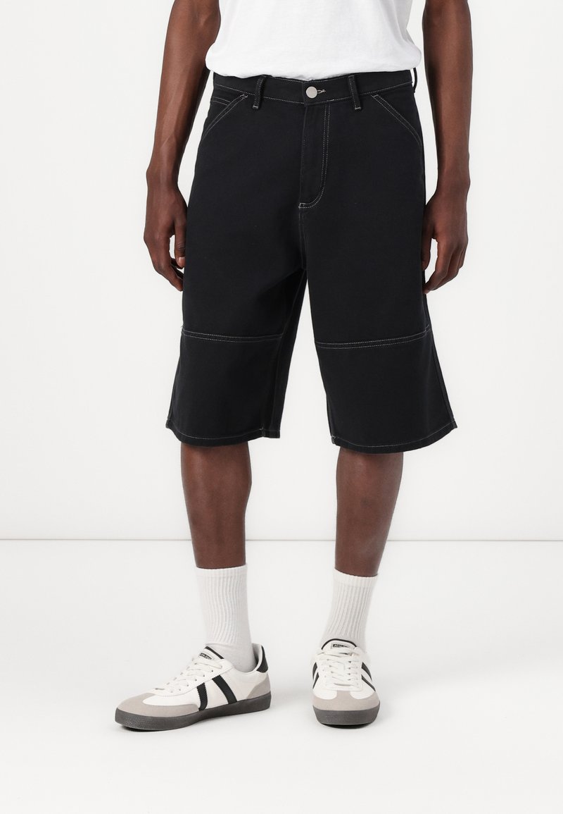 Svarta denimshorts med kontrasterande vita sömmar, rak skärning och standardmidja, tillsammans med vita strumpor och sportskor.