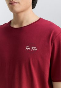 Burgundy kortærmet t-shirt lavet af blødt stof, med rund halsudskæring og et cirkulært logo med teksten "Tom Tailor" i hvid.