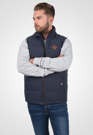 Uomo con la barba che indossa un gilet trapuntato senza maniche blu navy sopra una felpa grigia, in piedi con le braccia incrociate, guardando di lato.