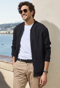 Black textured bomber jacket over a white t-shirt, beige pants, black belt, and a watch. Background features a coastal view.