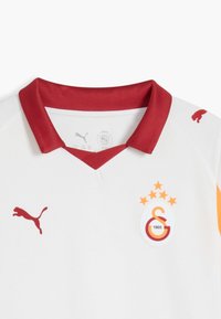 Camiseta deportiva blanca con un cuello rojo, que presenta un logo de Puma rojo, un emblema de club con cinco estrellas y acentos naranjas en las mangas.