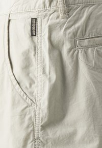 Gros plan sur un tissu beige avec deux poches et une petite étiquette de marque noire et orange portant le nom "Napapijri" cousue au-dessus de la poche gauche.