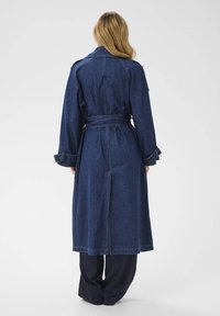 Jeanska trench jakna v temno modri barvi z pasom, dolgimi rokavi in stranskimi razporki. Ima velike reverje in ojačane šive.