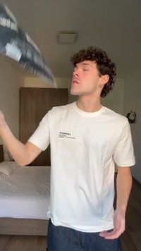 Jonge man die een zwarte tas of jas gooit in een slaapkamer met een bed en een kast op de achtergrond.