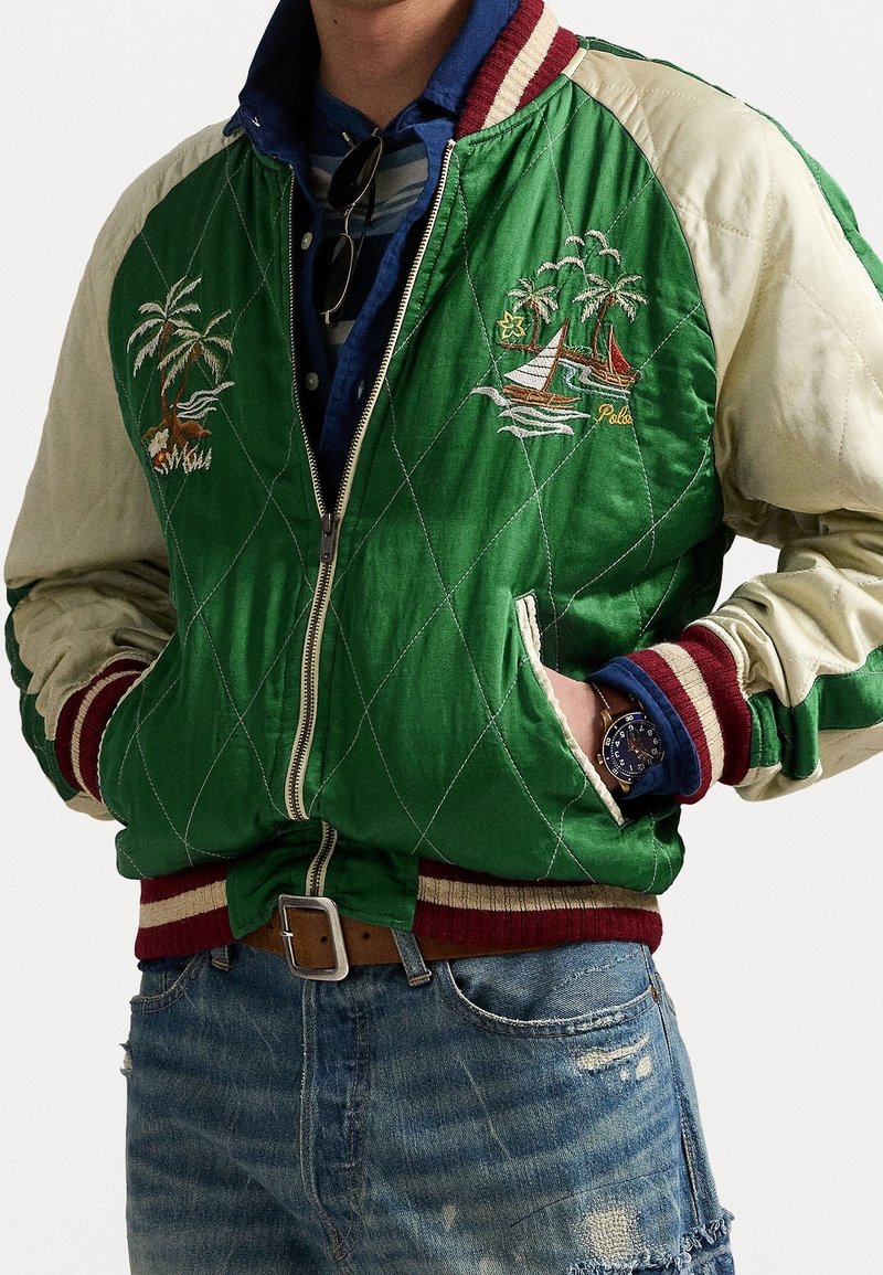 Personne portant une veste matelassée en satin vert et crème avec des bateaux à voile et des palmiers brodés, des poignets rayés, une montre bleue et un jean usé.
