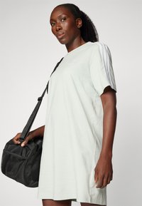 Robe t-shirt oversize vert clair à manches courtes, avec des rayures blanches sur une épaule et un petit logo. Sac noir avec des détails en filet.