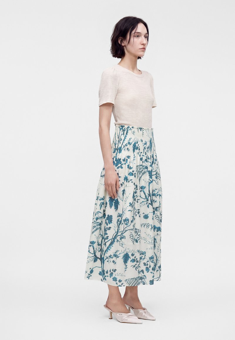 Frau steht, trägt ein beiges kurzärmeliges Oberteil und einen weißen langen Rock mit blauem Blumenmuster sowie hellrosa, hochhackige Mules.