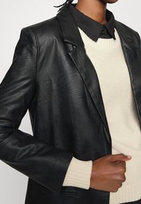 Svart läderblazer med en strukturerad design, som har slagkrage, och en mjuk krämfärgad stickad tröja under. Synliga ribbade manschetter.