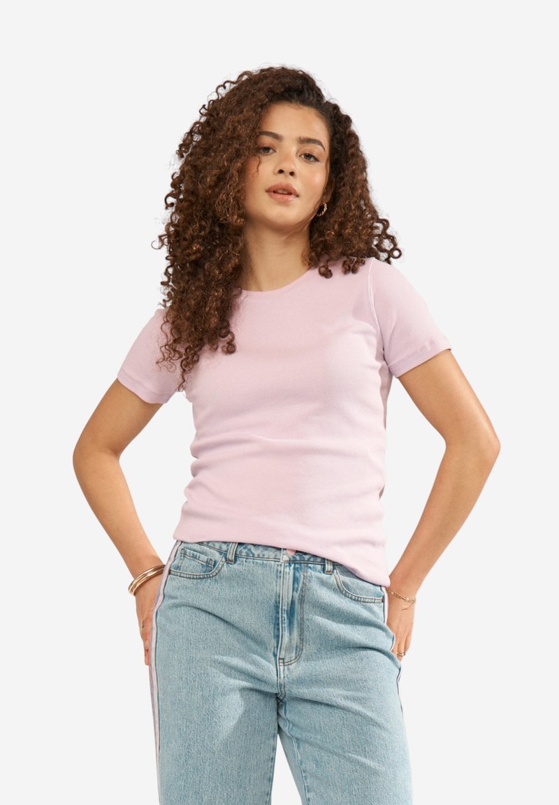 Femme aux cheveux bouclés portant un t-shirt rose clair à manches courtes et un jean taille haute bleu clair, debout les mains sur les hanches.