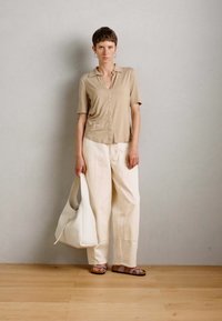 Woman standing against gray wall wearing beige short-sleeve buttoned shirt, white wide pants, sandals, and holding a large white bag.