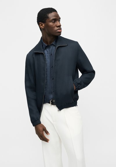 Homme portant une veste zippée bleu marine par-dessus une chemise à motif sombre et un pantalon blanc, debout avec une main dans la poche, regardant sur le côté.