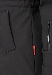Svart jacka med en zip-ficka, designade sidopaneler och kontrasterande röd etikett; slät textur med ett robust och funktionellt utseende.